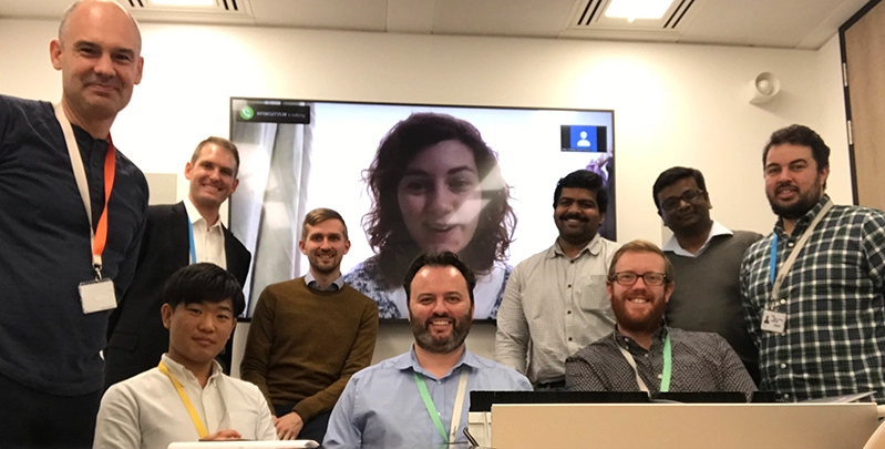 A group of nine men with name tags looking into the camera. Behind them is a woman on the screen connected virtually.