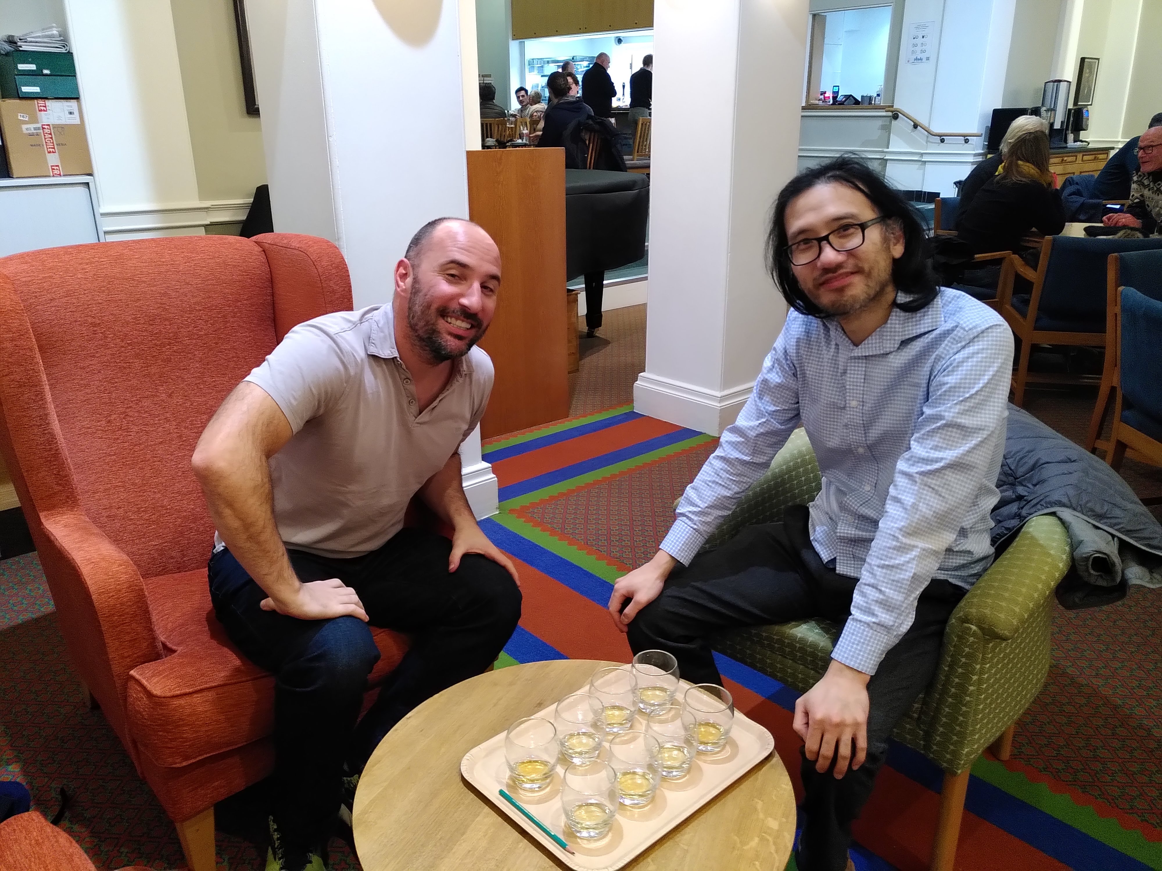 Two men sitting on two separate arm chairs with many whiskey glasses in the middle on a coffee table.